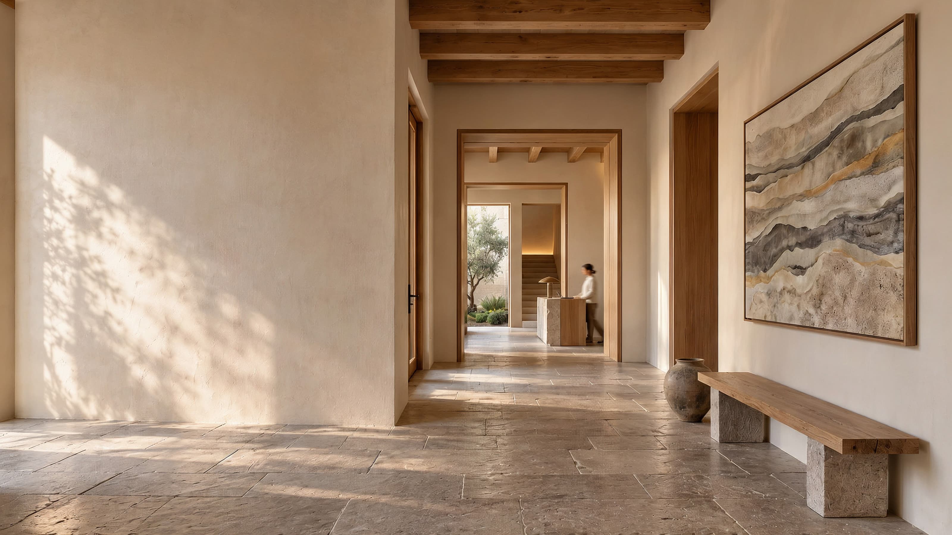 An arrival hall of lime plaster and limestone. Beyond a run of open doorways a guest passes a stone reception desk, and a large mineral-toned painting hangs above an oak bench.